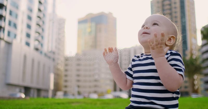 De zuigeling die van de babyjongen in de de zomerzitting lachen op het gras op de achtergrondgebouwen