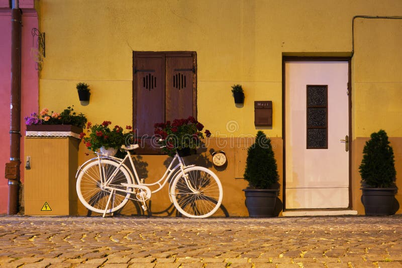 Decorative white bicycle with flower baskets on it, a clock on wall nearby, a locked white door, and a closed window.