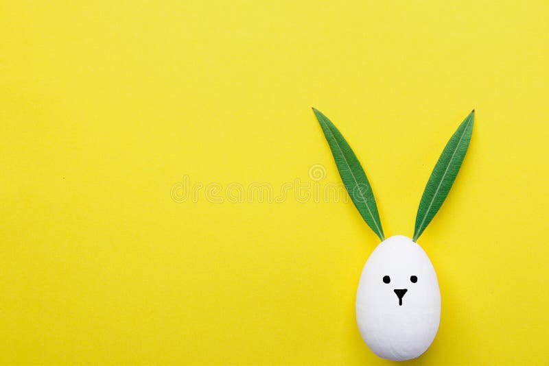 Decorative White Painted Easter Egg Bunny with Drawn Cute Kawaii Face. Green Leaves as Ears. Pastel Yellow Background. Spring Holiday Crafts Kids Concept. Greeting Card Poster Banner. Copy Space