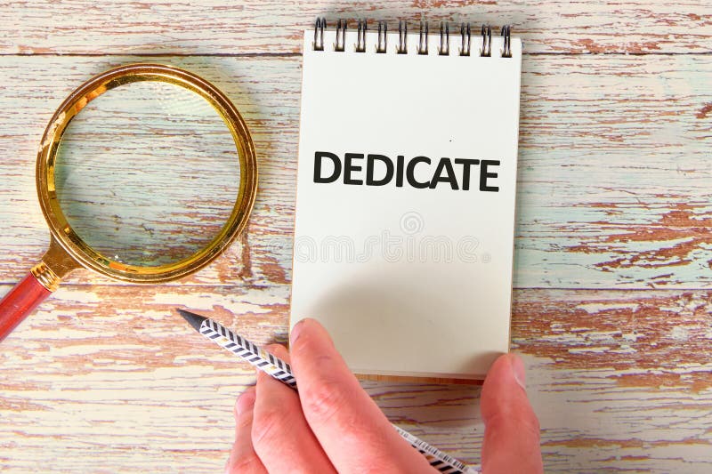 DEDICATE word written on a blank sheet of notepad in a composition with a man's hand and a magnifying glass royalty free stock image