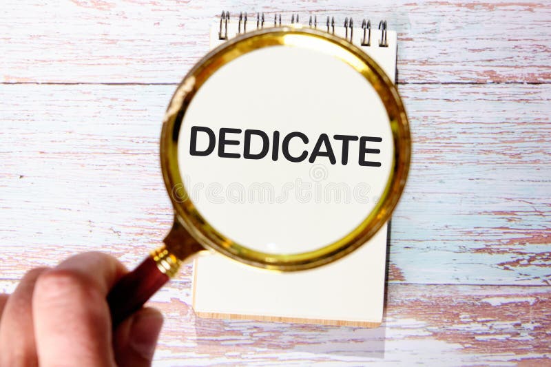 DEDICATE word written through a magnifying glass in a notebook against the background of old boards stock image