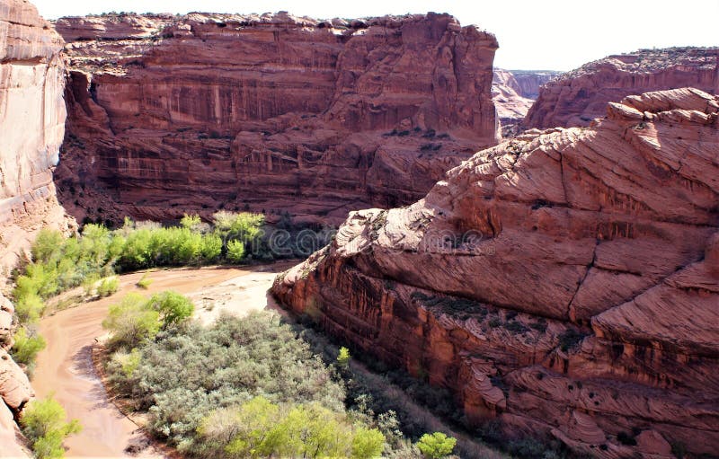 A deep blue cloudless sky contrasts sharply with the ochre red of the lithified sand grain bedrock. Sweeping patterns delineate the steep rocky slopes. Shallow river flows slowly at base of cliff with pueblos. A deep blue cloudless sky contrasts sharply with the ochre red of the lithified sand grain bedrock. Sweeping patterns delineate the steep rocky slopes. Shallow river flows slowly at base of cliff with pueblos.