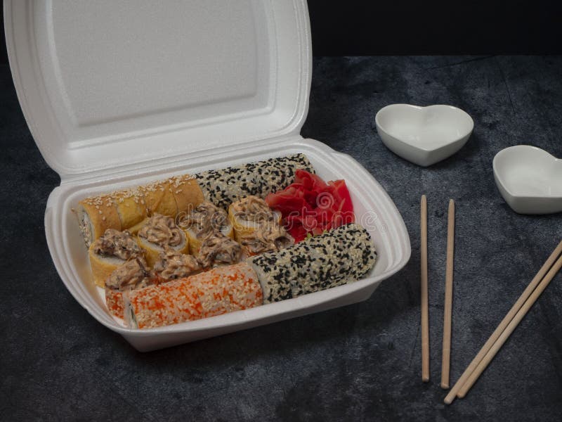 Defferent type of rolls or sushi set in a white plastic take-away box with chopsticks and empty bowls for sauce and ginger