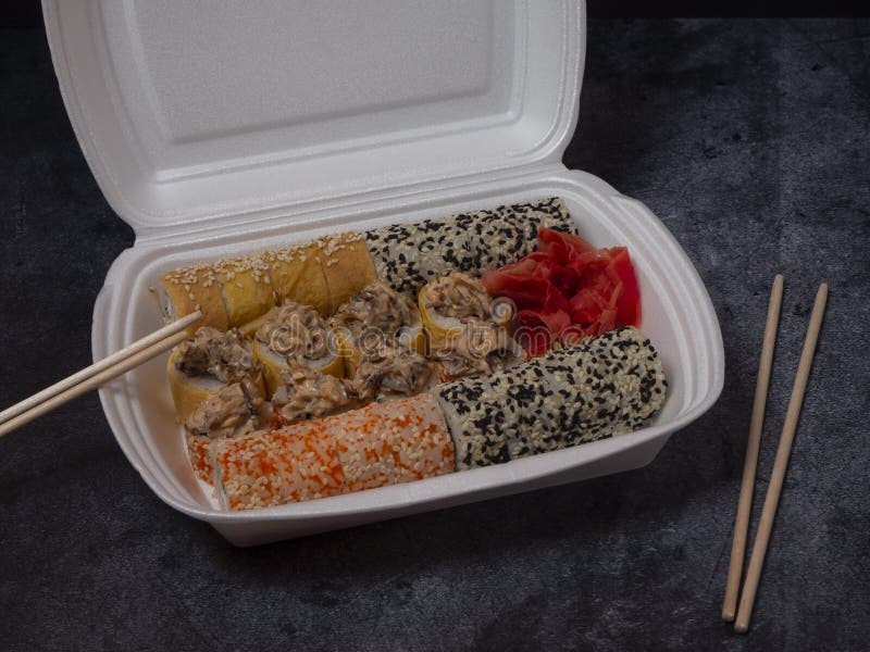 Defferent type of rolls or sushi set in a white plastic take-away box with chopsticks on gray stone table.
