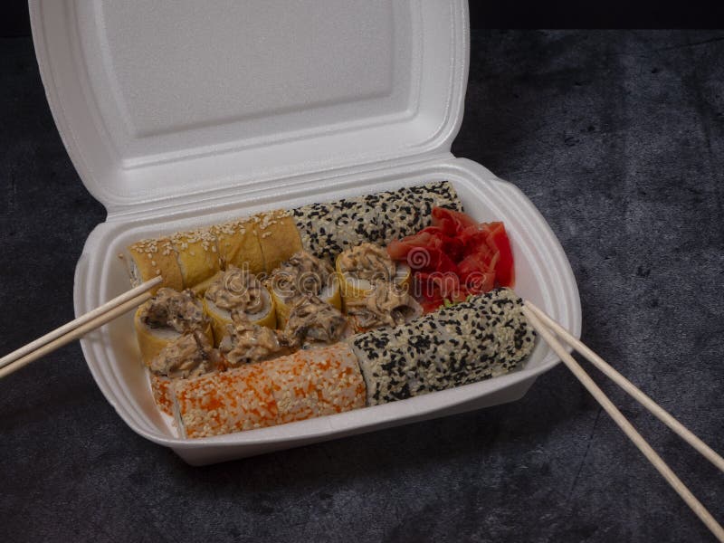 Defferent type of rolls or sushi set in a white plastic take-away box with chopsticks on gray stone table.