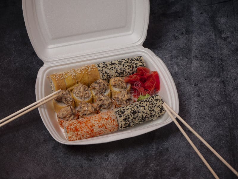 Defferent type of rolls or sushi set in a white plastic take-away box with chopsticks on gray stone table.