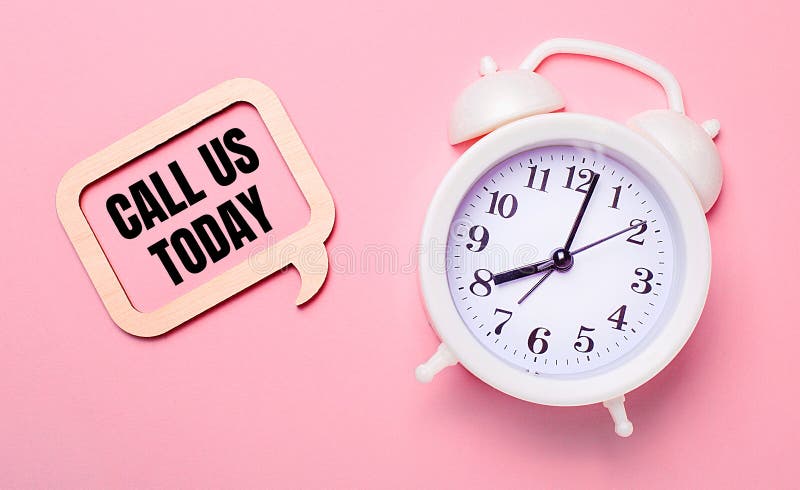 On a delicate pink background, a white alarm clock and a wooden frame with the text CALL US TODAY royalty free stock photo