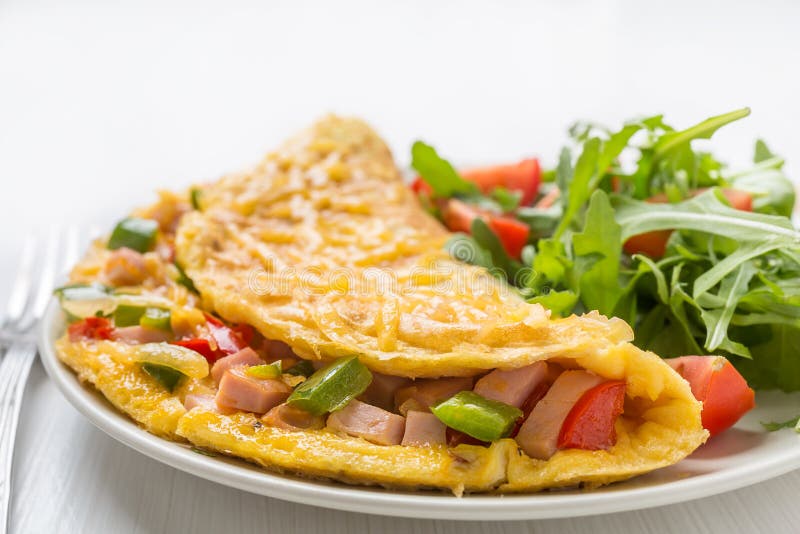 Delicious Egg Omelette with Cheese, Ham and Vegetables on a Plate served with Rucola and Tomatoes Salad on White Background. Closeup. Delicious Egg Omelette with Cheese, Ham and Vegetables on a Plate served with Rucola and Tomatoes Salad on White Background. Closeup.