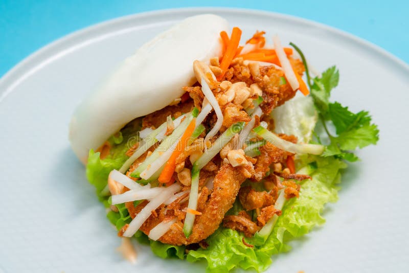 Delicious steamed bao bun on a plate with fried chicken and fresh vegetables - salad, carrots and roasted peanuts. Popular street