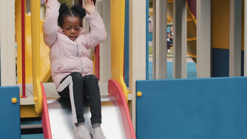 Den afrikanska amerikanska flickan spelar ensam på lekplatsens barnklättringssteg och glider ner på hoppande barn fritiden skolfli