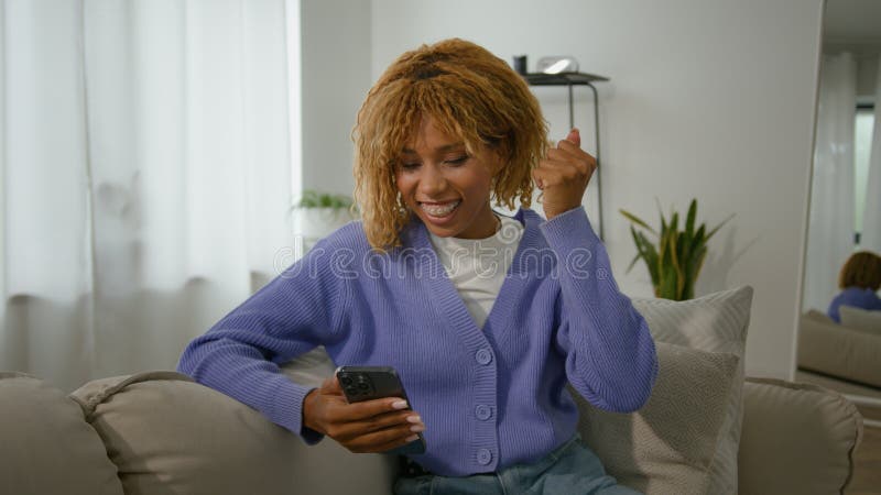 African American ethnic girl woman lady relax on couch at home with mobile phone win game online betting victory has discount in net virtual winning e-commerce triumph celebrate achieve smartphone. High quality 4k footage. African American ethnic girl woman lady relax on couch at home with mobile phone win game online betting victory has discount in net virtual winning e-commerce triumph celebrate achieve smartphone. High quality 4k footage