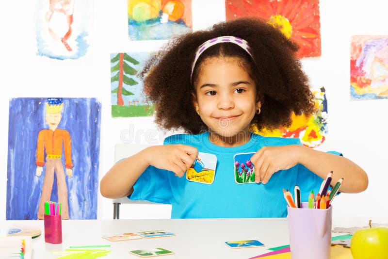 Cute African girl playing developmental game holding cards matching relation by table sitting in playroom with wall behind full of children drawings. Cute African girl playing developmental game holding cards matching relation by table sitting in playroom with wall behind full of children drawings