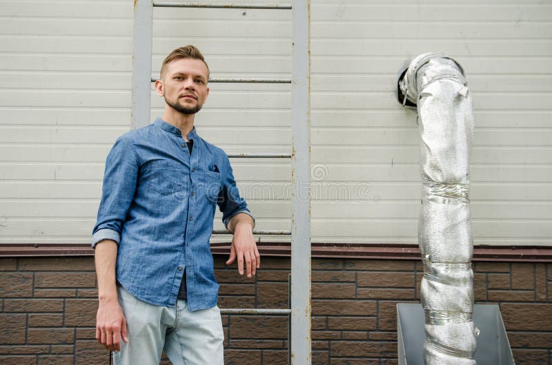 Den allvarliga skäggiga unga mannen i grov bomullstvillskjorta står nära väggen av industribyggnad nära stege och ser direkt på fotografering för bildbyråer Den allvarliga skäggiga unga mannen i grov bomullstvillskjorta står nära väggen av industribyggnad nära stege och ser direkt på fotografering för bildbyråer