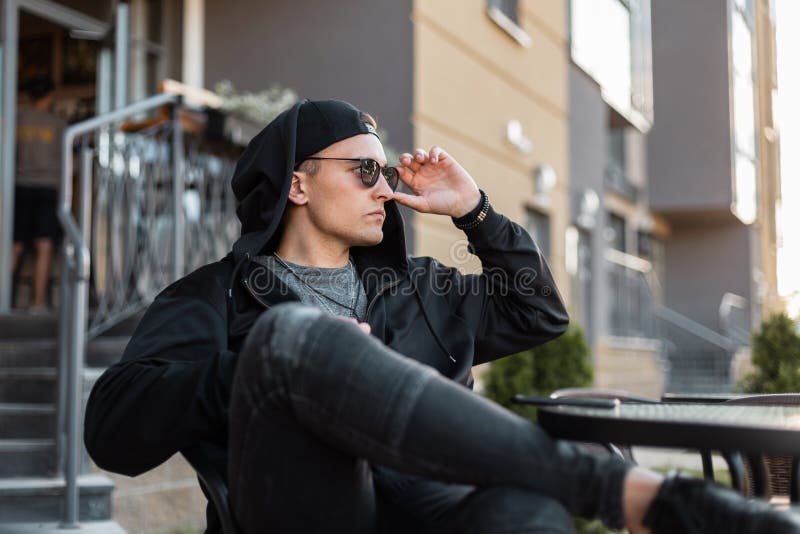 Den amerikanska unga hipstermannen i stilfull svart kläder i trendig solglasögon i en baseballmössa i jeans sitter i ett sommarka arkivbilder Den amerikanska unga hipstermannen i stilfull svart kläder i trendig solglasögon i en baseballmössa i jeans sitter i ett sommarka arkivbilder