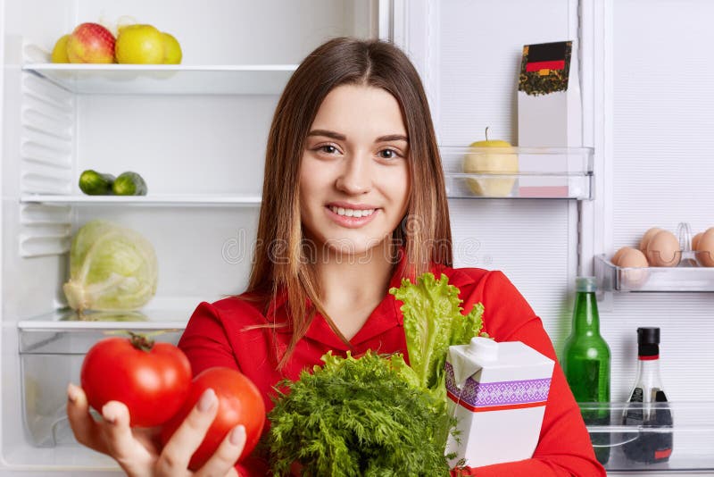 Pleasant looking female model with cheerful expression holds fresh vegetables which she bought in grocer`s shop, stands near opened fridge at kitchen. People, nutrition and vegan people concept. Pleasant looking female model with cheerful expression holds fresh vegetables which she bought in grocer`s shop, stands near opened fridge at kitchen. People, nutrition and vegan people concept