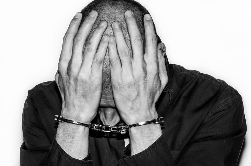 Den arresterade och handfängslade brottsliga mannen med handbojor på hans händer täcker hans huvud och beklagar i polisstationen royaltyfri fotografi Den arresterade och handfängslade brottsliga mannen med handbojor på hans händer täcker hans huvud och beklagar i polisstationen royaltyfri fotografi
