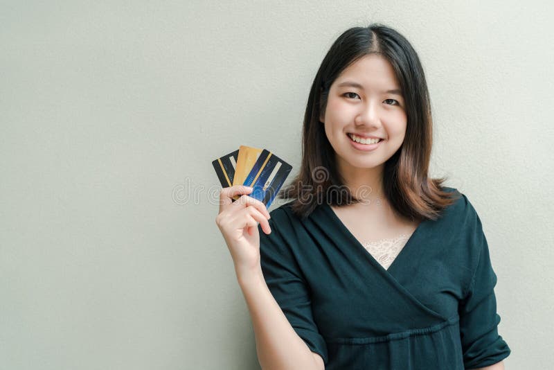 Den asiatiska härliga kvinnan som bär en svart skjorta, har en kreditkort i lyckligt framsidaanseende för hand i den gråa väggen arkivfoto Den asiatiska härliga kvinnan som bär en svart skjorta, har en kreditkort i lyckligt framsidaanseende för hand i den gråa väggen arkivfoto