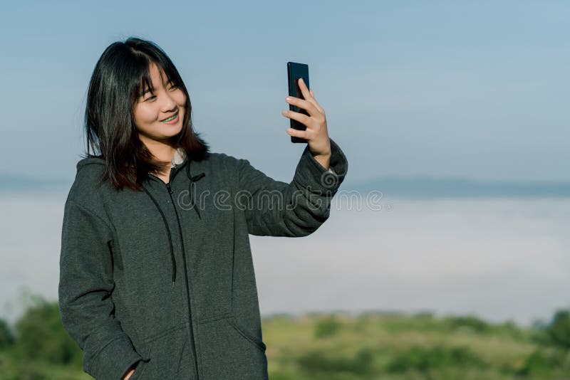 Den asiatiska kvinnan som bär ett vinteromslag, tar en selfietelefon, tar bilder i det turist- området bak dimman och bergen med royaltyfria bilder Den asiatiska kvinnan som bär ett vinteromslag, tar en selfietelefon, tar bilder i det turist- området bak dimman och bergen med royaltyfria bilder