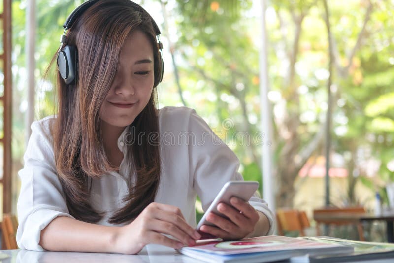Den asiatiska kvinnan som lyssnar till musik med den smarta telefonen för headphonen och för innehavet med mening lycklig och, ko royaltyfria foton Den asiatiska kvinnan som lyssnar till musik med den smarta telefonen för headphonen och för innehavet med mening lycklig och, ko royaltyfria foton