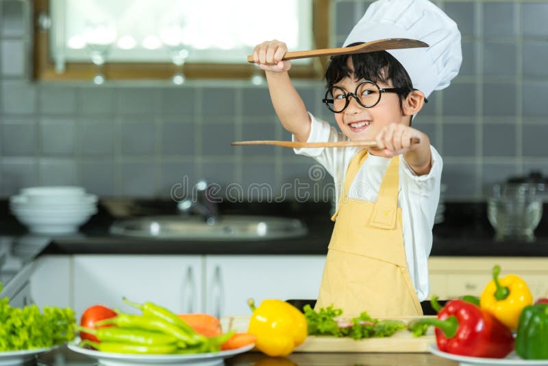 Den asiatiska lilla pojken kokar och skär grönsaker för att få en frisk sallad i köket, så glad och slappna av för att lära sig o royaltyfri fotografi