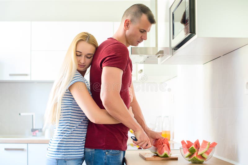 den attraktiva kvinnan kramar hennes make, medan han klipper den röda vattenmelon i deras stora moderna vita kök arkivfoto den attraktiva kvinnan kramar hennes make, medan han klipper den röda vattenmelon i deras stora moderna vita kök arkivfoto