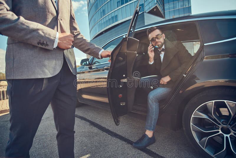 Den attraktiva mannen i solglas?gon talar vid smartphonen och sitter i bilen, medan hans assistent ?r den ?ppnande d?rren f?r hon arkivfoton Den attraktiva mannen i solglas?gon talar vid smartphonen och sitter i bilen, medan hans assistent ?r den ?ppnande d?rren f?r hon arkivfoton