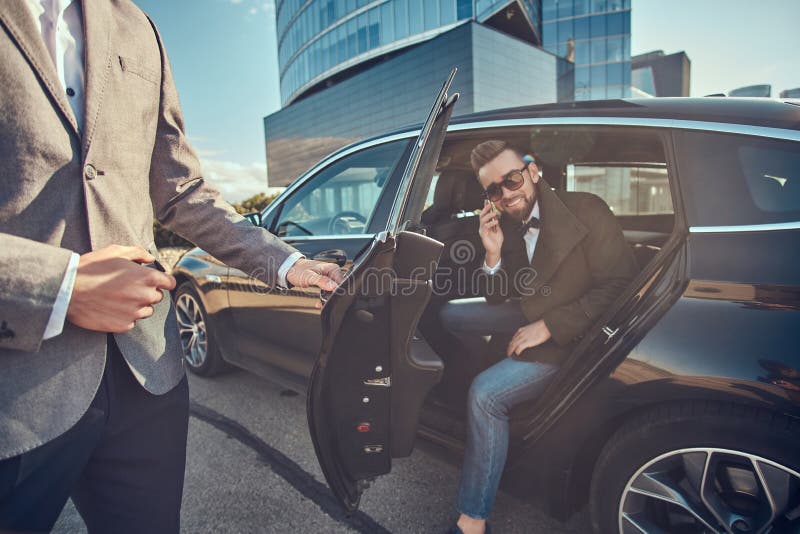 Den attraktiva mannen i solglas?gon talar vid smartphonen och sitter i bilen, medan hans assistent ?r den ?ppnande d?rren f?r hon royaltyfri bild Den attraktiva mannen i solglas?gon talar vid smartphonen och sitter i bilen, medan hans assistent ?r den ?ppnande d?rren f?r hon royaltyfri bild