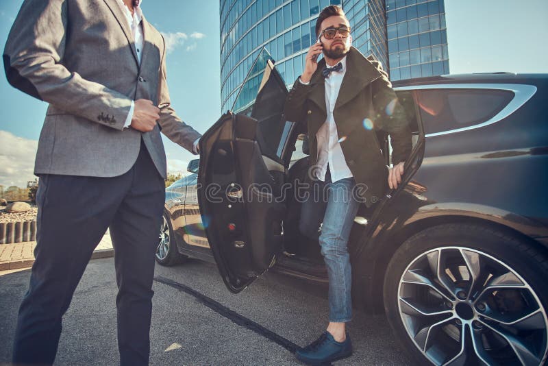 Den attraktiva mannen i solglas?gon talar vid smartphonen och sitter i bilen, medan hans assistent ?r den ?ppnande d?rren f?r hon royaltyfria foton Den attraktiva mannen i solglas?gon talar vid smartphonen och sitter i bilen, medan hans assistent ?r den ?ppnande d?rren f?r hon royaltyfria foton