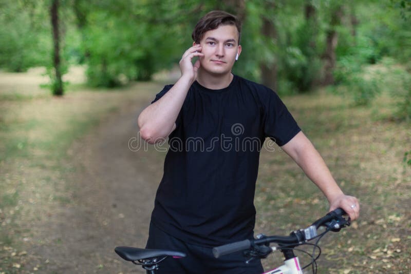Den attraktiva unga caucasian mannen trycker på hans vita hörlurar som rymmer cykeln som står på övergiven, parkerar arkivbilder Den attraktiva unga caucasian mannen trycker på hans vita hörlurar som rymmer cykeln som står på övergiven, parkerar arkivbilder