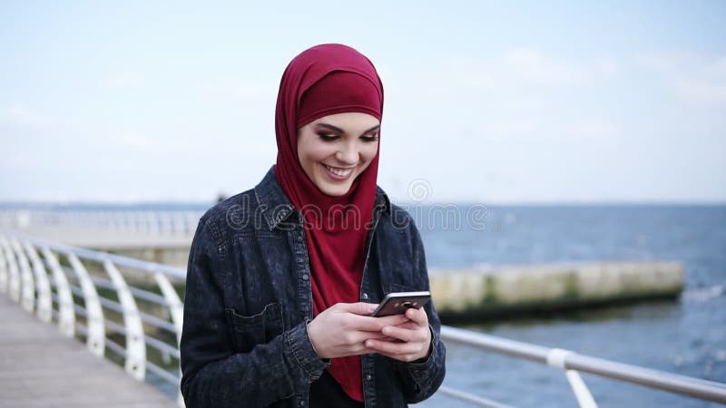 Den attraktiva unga flickan med hijab på hennes huvud ler, medan smsa till någon och bläddra något på henne
