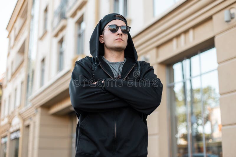Den attraktiva unga hipstermannen i en stilfull svart med huva tröja i ett trendigt lock i solglasögon poserar nära en tappningby royaltyfri fotografi Den attraktiva unga hipstermannen i en stilfull svart med huva tröja i ett trendigt lock i solglasögon poserar nära en tappningby royaltyfri fotografi