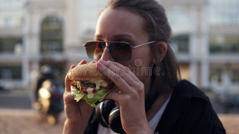 Den attraktiva unga kvinnan som glatt ler, den smakliga hamburgaren för håll räcker och äter itu, den Iklädd tillfällig dräkt, in