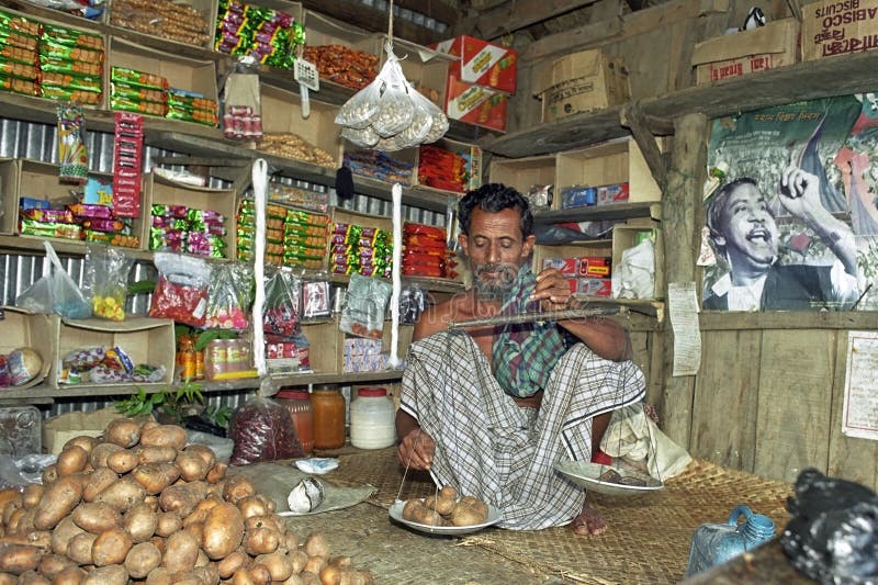 Bangladesh, Charburhan village on the island of Charkajal, Bay of Bengal: Portrait of Bengal man, tradesman, in the interior of his grocert shop. He is to weigh in squat potatoes. On the floor of this grocery store is a mountain potatoes and foods packed on the shelves. All wares are displayed on shelves. Bangladesh, Charburhan village on the island of Charkajal, Bay of Bengal: Portrait of Bengal man, tradesman, in the interior of his grocert shop. He is to weigh in squat potatoes. On the floor of this grocery store is a mountain potatoes and foods packed on the shelves. All wares are displayed on shelves.