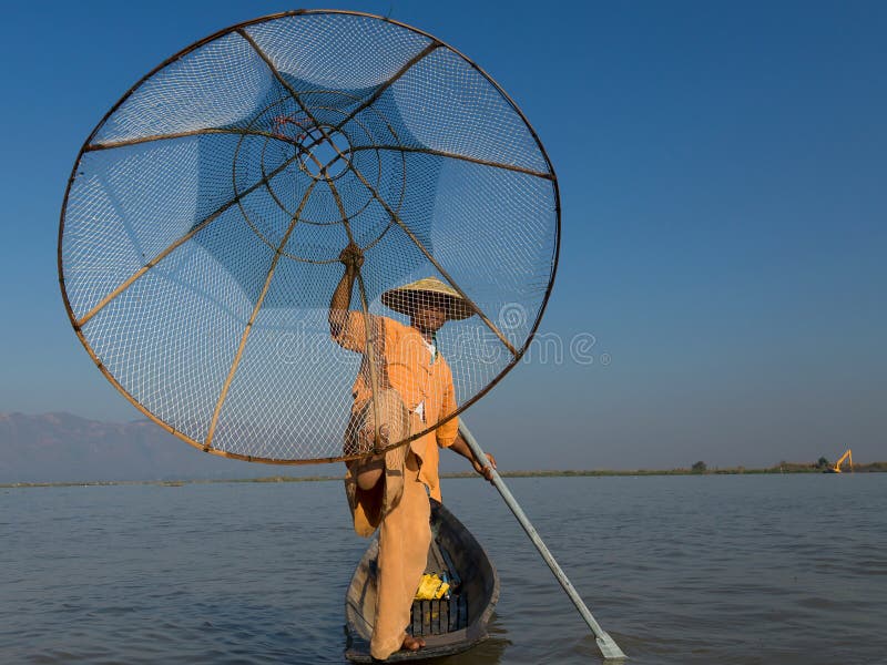 Den Burmese fiskarebenägenheten på åran som poserar i traditionell kläder med handen och foten som rymmer hans kotte, formade fis royaltyfria foton Den Burmese fiskarebenägenheten på åran som poserar i traditionell kläder med handen och foten som rymmer hans kotte, formade fis royaltyfria foton