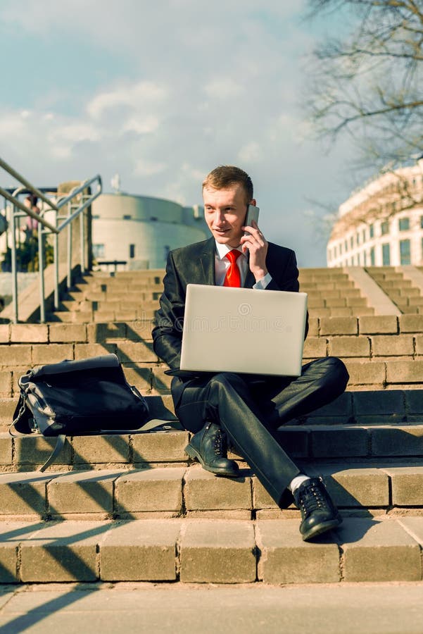 Den Businessan freelanceren arbetar i den öppna luften i staden En man i en svart dräkt och ett rött band sitter på momenten royaltyfri fotografi Den Businessan freelanceren arbetar i den öppna luften i staden En man i en svart dräkt och ett rött band sitter på momenten royaltyfri fotografi