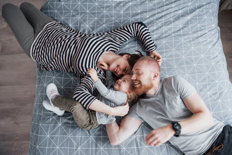 Den bästa sikten av härligt barn fostrar, fadern och deras dotter som ser kameran och ler, medan ligga på sänghuvudet till royaltyfri foto Den bästa sikten av härligt barn fostrar, fadern och deras dotter som ser kameran och ler, medan ligga på sänghuvudet till royaltyfri foto