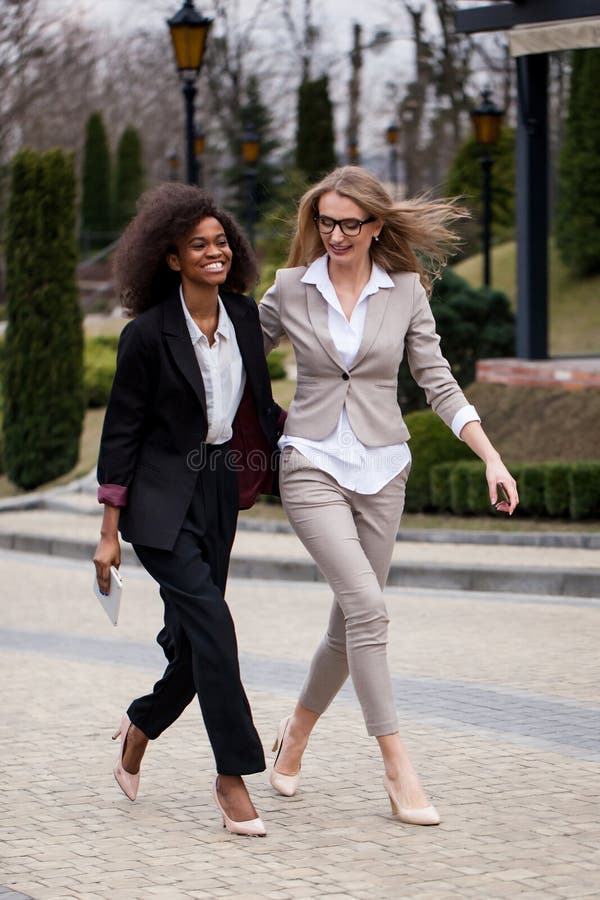 Den charmiga afrikanen och nätta caucasian affärskvinnor är skratta och le, medan promenera parkera royaltyfria foton Den charmiga afrikanen och nätta caucasian affärskvinnor är skratta och le, medan promenera parkera royaltyfria foton