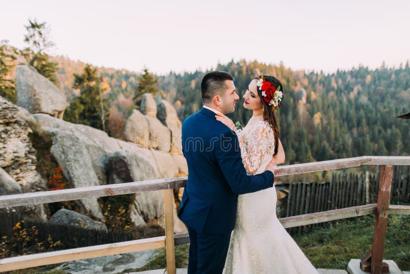 Den eleganta brudgummen i stilfulla blått passar rymma lovingly hans charmig vit klädde brudanseende på träplattformen med royaltyfri fotografi Den eleganta brudgummen i stilfulla blått passar rymma lovingly hans charmig vit klädde brudanseende på träplattformen med royaltyfri fotografi