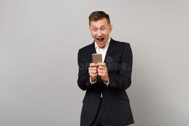 Surprised happy young business man screaming keeping mouth open wide, using mobile phone typing sms message isolated on grey background. Achievement career wealth business concept. Mock up copy space. Surprised happy young business man screaming keeping mouth open wide, using mobile phone typing sms message isolated on grey background. Achievement career wealth business concept. Mock up copy space