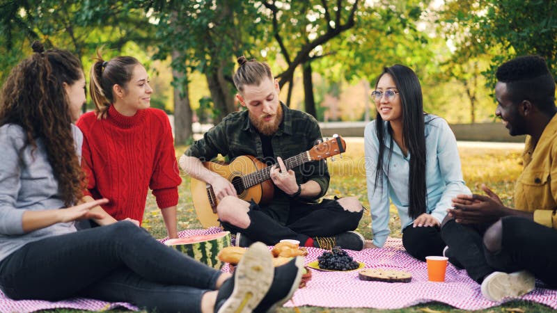 Den glade hipsteren är sjungande, och spela gitarrsammanträdet på filten parkera in med vänner, och att ha gyckel, folk är royaltyfria foton Den glade hipsteren är sjungande, och spela gitarrsammanträdet på filten parkera in med vänner, och att ha gyckel, folk är royaltyfria foton