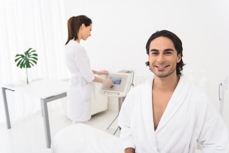 I like caring of my skin. Joyful young men is sitting in beautician office and smiling. Woman is standing and working with ultrasound equipment on background. I like caring of my skin. Joyful young men is sitting in beautician office and smiling. Woman is standing and working with ultrasound equipment on background