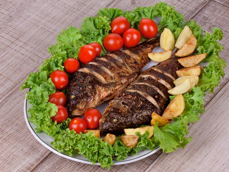 Den grillade fiskkarpen tjänade som med potatisar, tomater körsbär och sallad arkivbild Den grillade fiskkarpen tjänade som med potatisar, tomater körsbär och sallad arkivbild