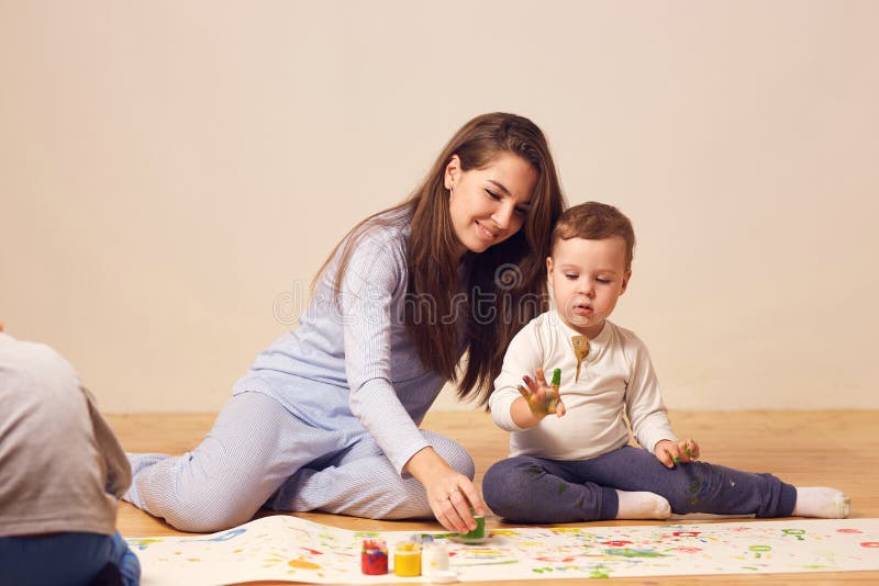 Den h?rliga lyckliga unga modern och hennes ikl?dda hem- kl?der f?r liten son sitter p? tr?golvet i rummet och royaltyfri bild Den h?rliga lyckliga unga modern och hennes ikl?dda hem- kl?der f?r liten son sitter p? tr?golvet i rummet och royaltyfri bild