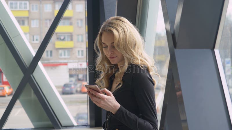 Den härliga blonda flickan nära ett ljust fönster tycker om telefonen och ringer ett meddelande arkivfoton Den härliga blonda flickan nära ett ljust fönster tycker om telefonen och ringer ett meddelande arkivfoton