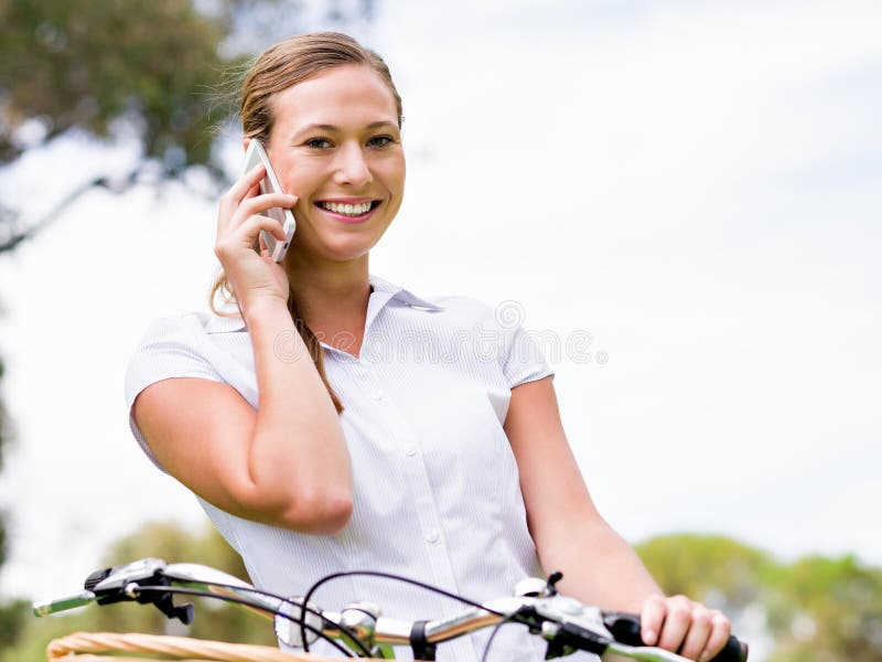 Den härliga unga blonda kvinnan med cykeln parkerar in samtal över telefonen royaltyfri foto Den härliga unga blonda kvinnan med cykeln parkerar in samtal över telefonen royaltyfri foto