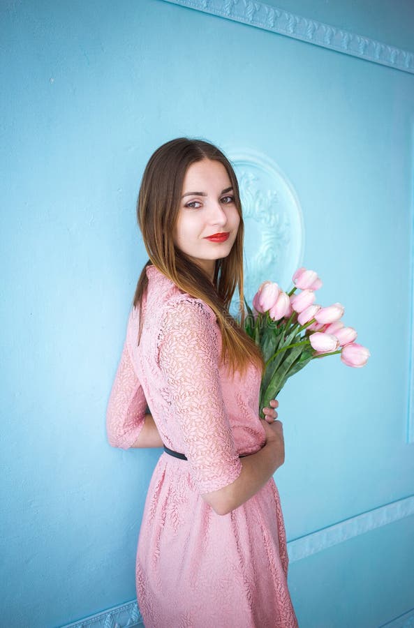 Den härliga unga kvinnan i rosa klänninginnehav i händer fjädrar tulpanblommabuketten på blå väggbakgrund royaltyfri bild Den härliga unga kvinnan i rosa klänninginnehav i händer fjädrar tulpanblommabuketten på blå väggbakgrund royaltyfri bild