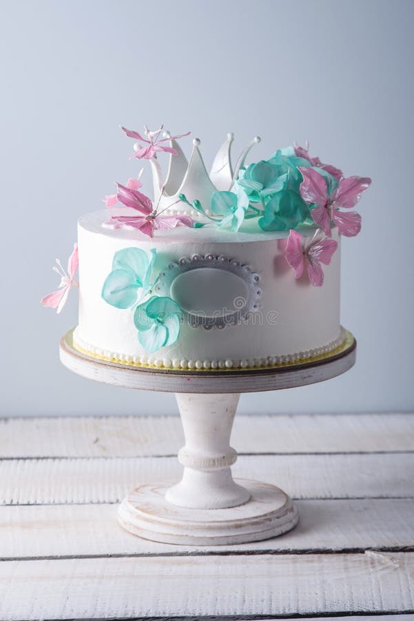 Den härliga vita kakan som dekoreras med rosa färger och turkosblommor, och en prinsessa krönar Begrepp av efterrätter för flicko royaltyfri fotografi Den härliga vita kakan som dekoreras med rosa färger och turkosblommor, och en prinsessa krönar Begrepp av efterrätter för flicko royaltyfri fotografi