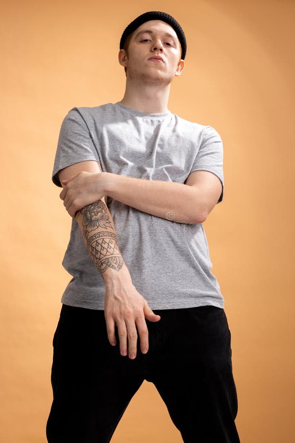 Den iklädda kaxiga stilfulla grabben en grå skjorta, en svart jeans och en svart hatt med tatueringen på hans hand står på beiga royaltyfri foto Den iklädda kaxiga stilfulla grabben en grå skjorta, en svart jeans och en svart hatt med tatueringen på hans hand står på beiga royaltyfri foto