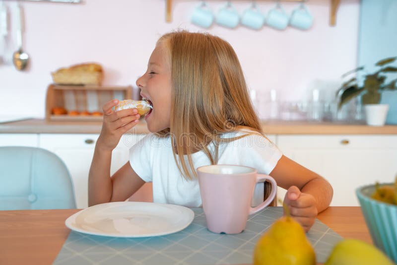Den lilla flickan äter frukost hemma på morgonen och njuter av att äta te god mat royaltyfri bild Den lilla flickan äter frukost hemma på morgonen och njuter av att äta te god mat royaltyfri bild