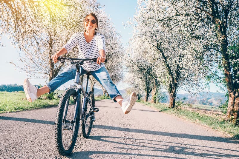 Den lyckliga le kvinnan fördelar glatt ben på cykeln på landsvägen under blomningträd Våren är den kommande begreppsbilden arkivfoto